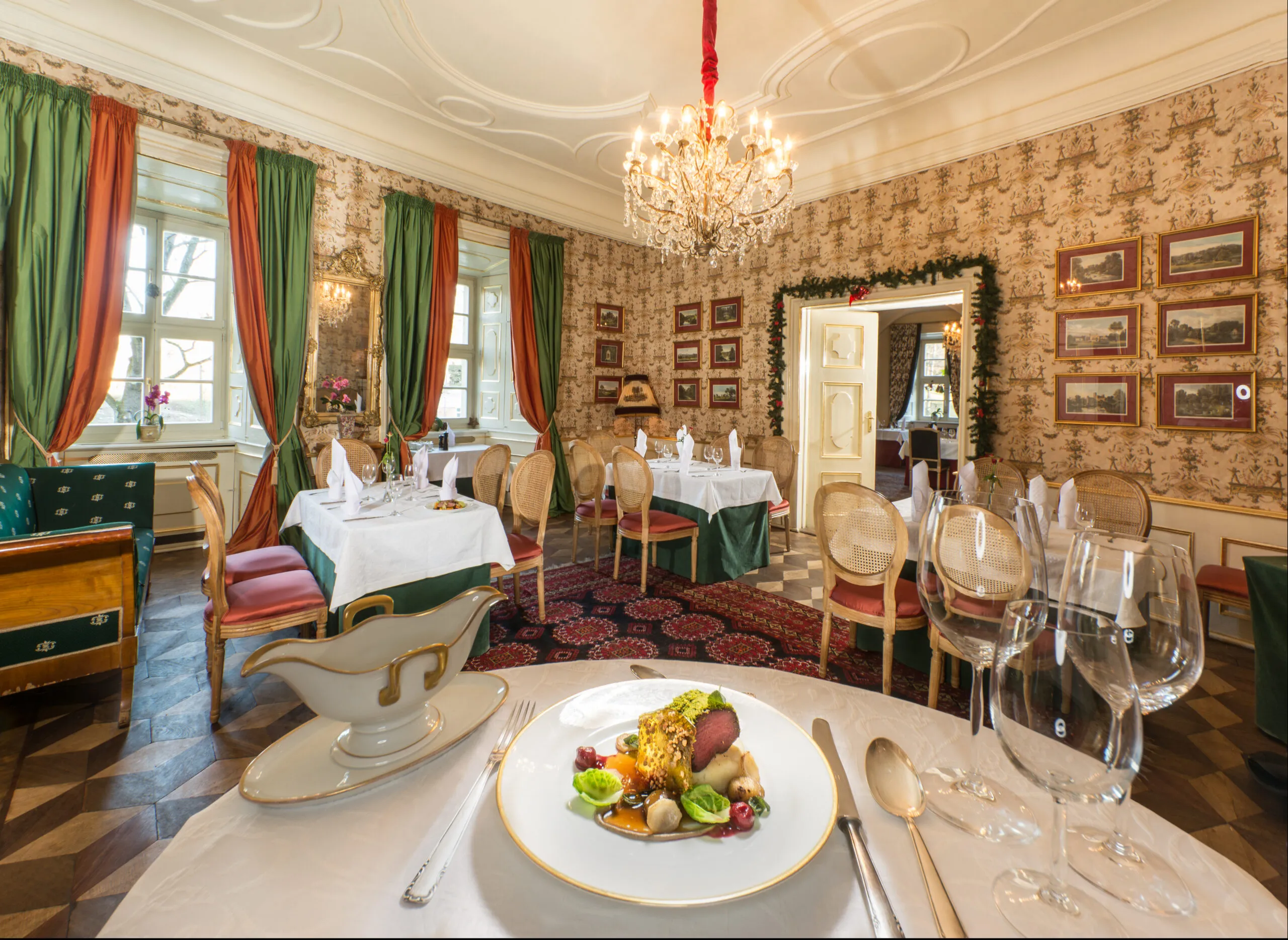 Wnętrze restauracji w stylu historyzującym, Na oknach duże, kotarowe zasłony. Stoły zastawione, przyszykowane na uroczystość. Na ścianach wzorzyste tapety oraz dużo zdjęć w ramach. Drewniany parkiet, przykryty prostokątnym dywanem we wzór geometryczny. Na pierwszym planie talerz z wykwintnym posiłkiem.