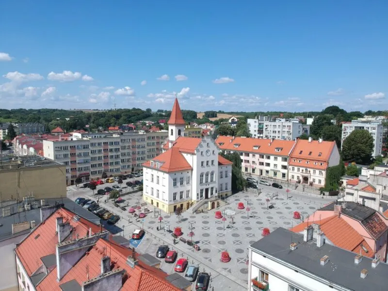 Zdjęcie przedstawia rynek Trzebnicy z centralnie ustawionym ratuszem z wysoką wieżą i czerwonym dachem. Przy głównym placu widać ustawione samochody.