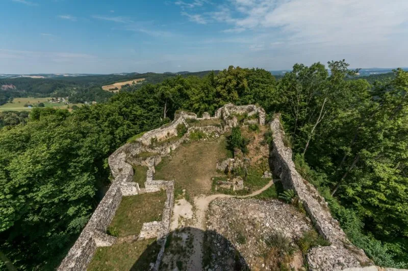 Widok znad murów kamiennego zamku na tle błękitnego nieba. Dookoła wiele drzew i krzewów.