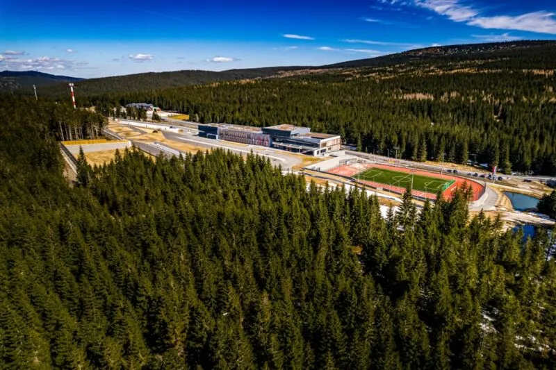 Duży ośrodek sportowy zlokalizowany w środku lasu. Widoczne boisko sportowe oraz bieżnia.