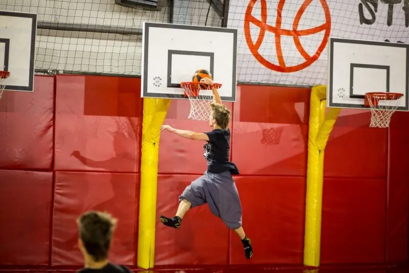 Młody mężczyzna wyskakuje wysoko do kosza na trampolinie w hali Jump 4U we Wrocławiu, a drugi uczestnik obserwuje próbę z dołu.