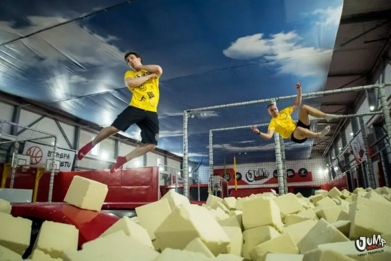 Dwie osoby w żółtych koszulkach skaczą jednocześnie nad dużym basenem z kolorowymi gąbkami w jasno oświetlonej hali parku trampolin Jump 4U.