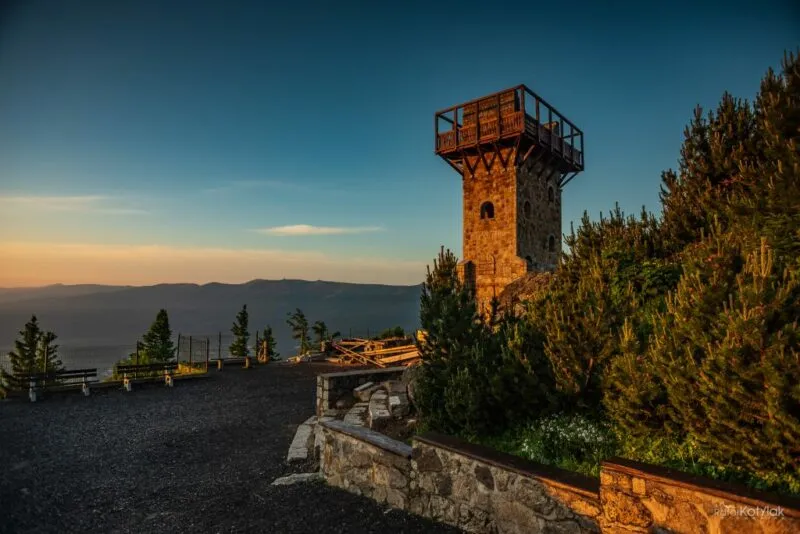 Kamienna wieża, z platformą widokową, wyłaniająca się zza krzewów. W tle panorama gór. Zachodża