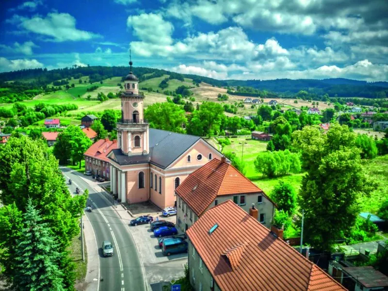Widok na zabytkowy kościół przy ulicy. Widok na panoramę okolicy Głuszycy Górnej z pofałdowanymi, średniozalesionymi wzniesieniami w tle.