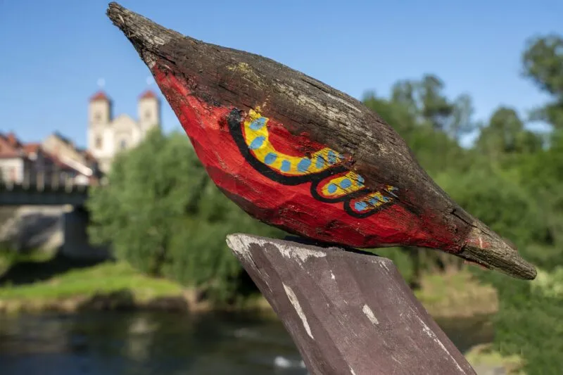 A wooden sculpture of a bird painted red, blue, and yellow stands on a balustrade in Bardo, with a blurred church and greenery in the background.