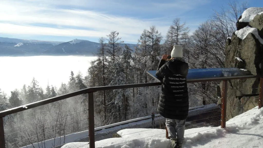 Zimowy widok z niezadaszonego tarasu widokowego znajdującego się na skraju góry z widocznymi w oddali Karkonoszami. Na pierwszym planie fotografująca widok turystka.