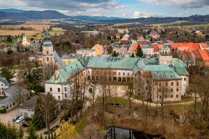 Widok z lotu ptaka na zamek Międzylesie ukazuje malowniczy kompleks historycznych budowli z czerwonymi dachami i wieżami, otoczony zielenią i wpisany harmonijnie w krajobraz górskiej doliny.