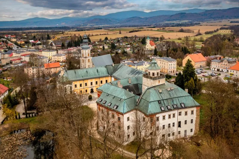Widok z lotu ptaka na zamek Międzylesie ukazuje malowniczy kompleks historycznych budowli z czerwonymi dachami i wieżami, otoczony zielenią i wpisany harmonijnie w krajobraz górskiej doliny.