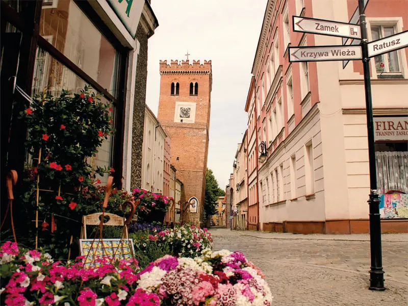 Widok z poziomu ulicy na krzywą, ceglaną nieotynkowaną wieżę.