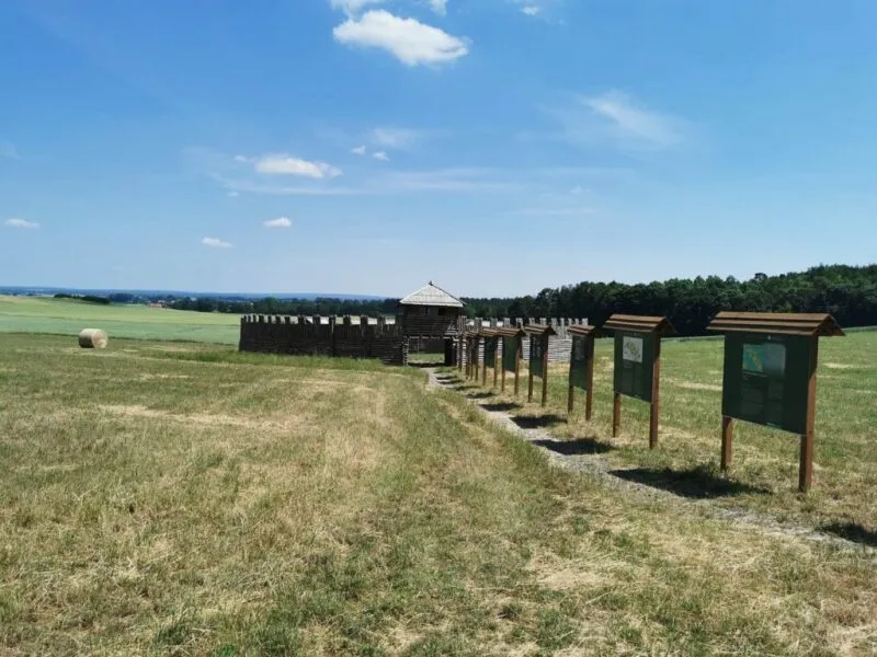 Rząd tablic edukacyjnych prowadzi przez trawiasty teren w Obiszowie do drewnianej palisady i wieży repliki średniowiecznego grodu.