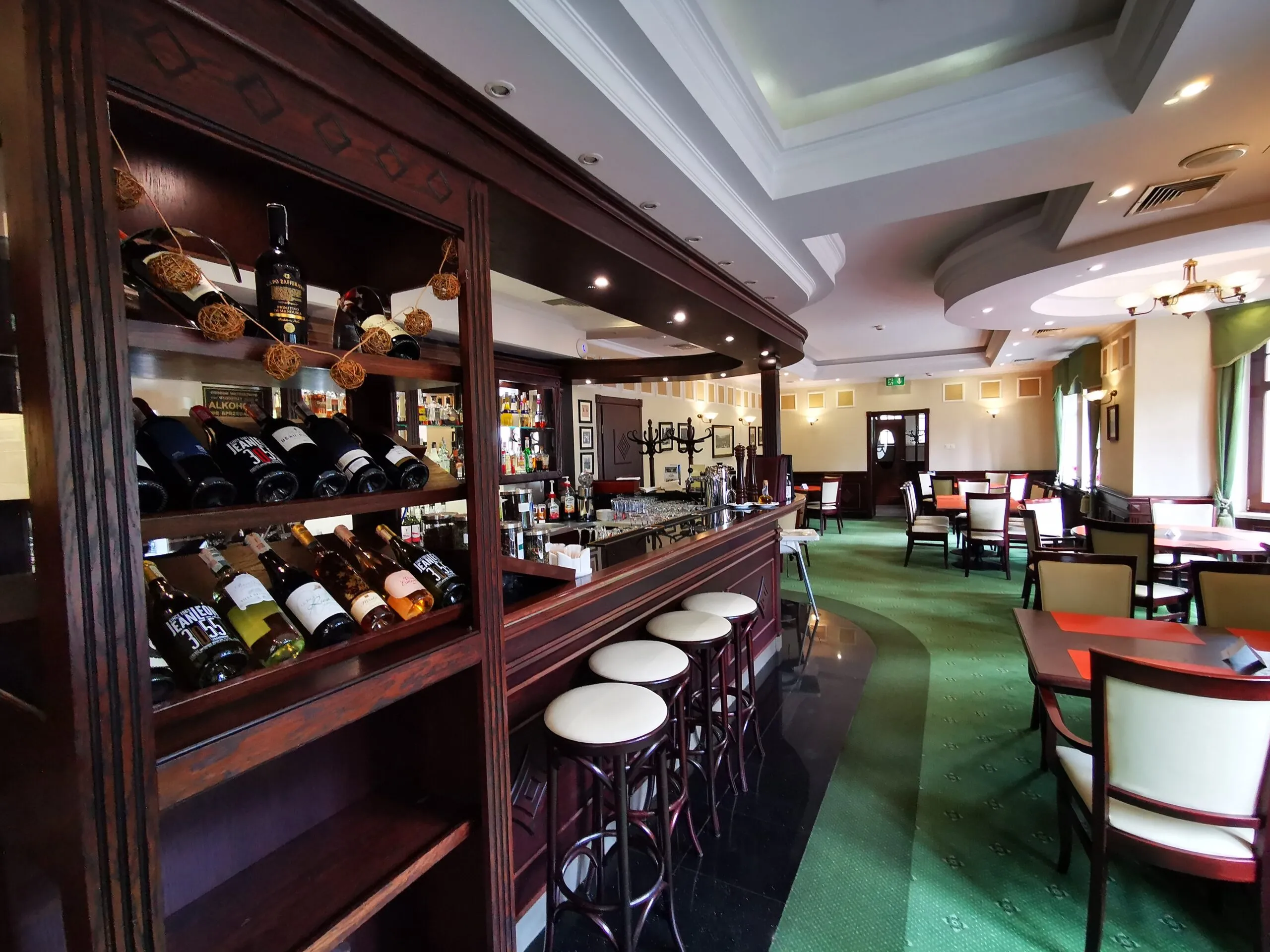 The hotel bar made of dark wood features shelves with bottles, high stools, and rows of tables in the cozy interior of the Bukowy Park Hotel.