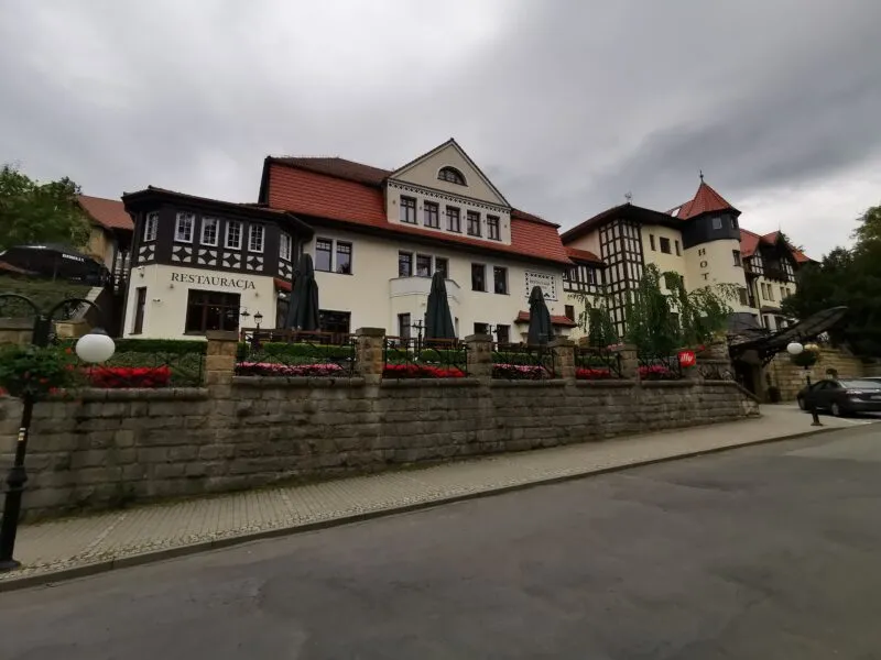 Světlá budova hotelu Bukowy Park v Polanici-Zdroji s tmavými dřevěnými štíty a červenou střechou stojí u ulice za kamennou zídkou.