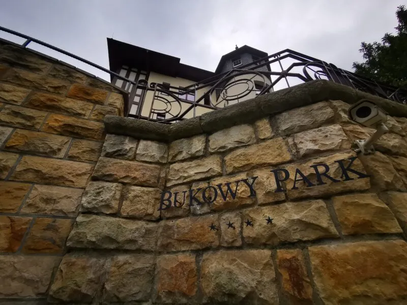 Kamennou zeď u hotelu Bukowy Park v Polanici-Zdroji zdobí kovové zábradlí a vyrytý název objektu, viditelný na pozadí zatažené oblohy.