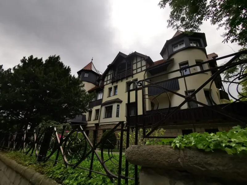 Hotel Bukowy Park w Polanicy-Zdroju ma jasną elewację z ciemnymi drewnianymi detalami, balkonami i wieżyczkami, otoczoną mokrą zielenią ogrodu.