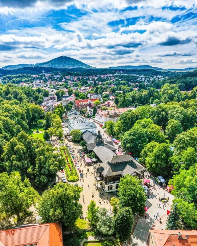 Zdjęcie z drona przedstawiające park zdrojowy latem wraz z drewnianym, bogato zdobionym budynkiem zabytkowej pijalni wody, a także widokiem na pobliskie góry.