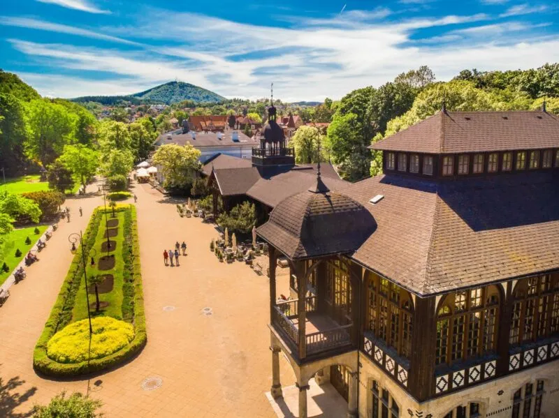 Zdjęcie z drona przedstawiające park zdrojowy latem wraz z drewnianym, bogato zdobionym budynek zabytkowej pijalni wody. 
