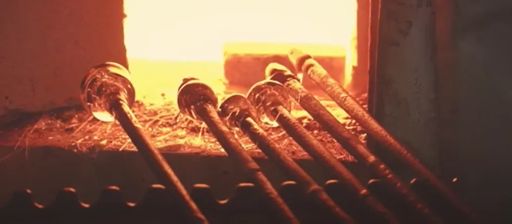 Rozgrzane szklane pręty lśnią pomarańczowym światłem przy otwartym piecu Huty Szkła Julia, ukazując moment ręcznego formowania szkła.