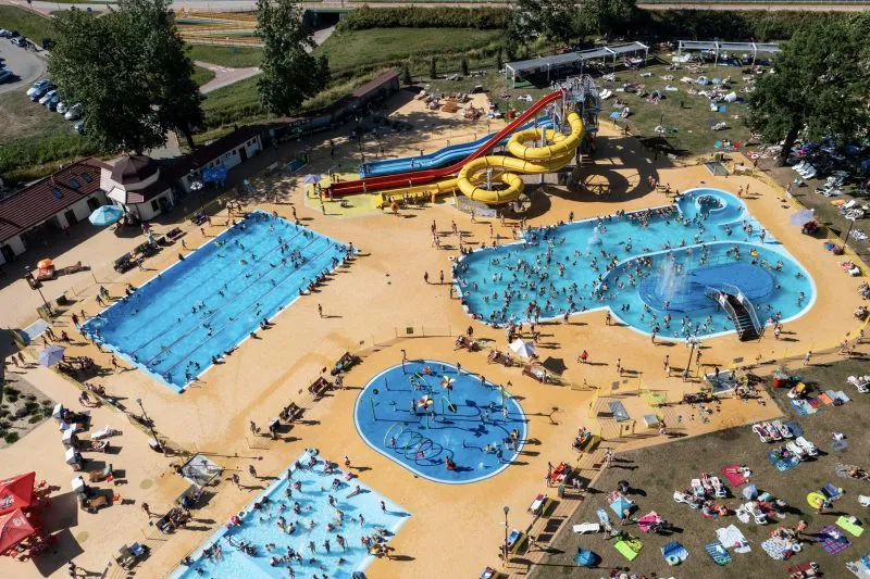 Widok z lotu ptaka na rozległy kompleks Aquafun z kilkoma basenami, kolorowymi zjeżdżalniami, plażą i licznymi gośćmi odpoczywającymi na trawie.