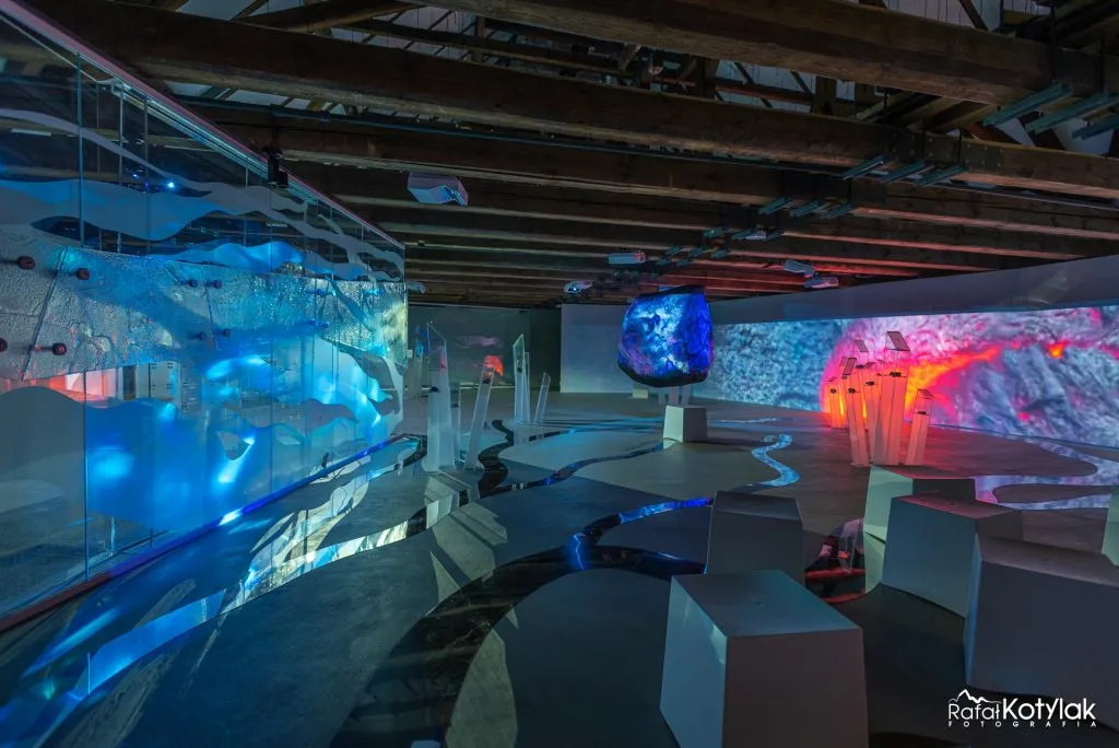 The multimedia hall of the Nature and Education Center in Sobieszów features irregular platforms, projections of rocks and water, and exhibits illuminated in blue and red.