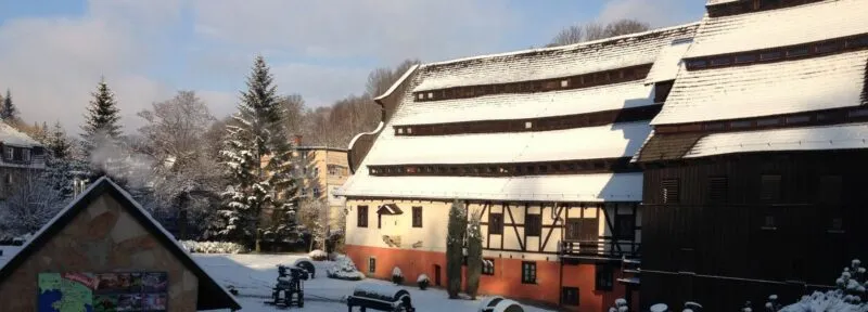 Zabytkowy budynek młynu papierniczego z czarnym dachem oraz beżowo-łososiową elewacją. Na dachu, krzewach i ziemi leży niewielka warstwa śniegu.