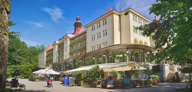 Słoneczny widok z poziomu ulicy na budynek uzdrowiska z widocznymi ogródkami gastronomicznymi.