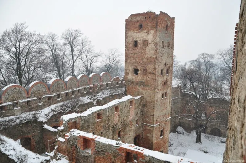 widok z górnej części obiektu na boczną wieżę zamku. Na drugim planie znajdują się drzewa a cały teren z obrazu jest ośnieżony