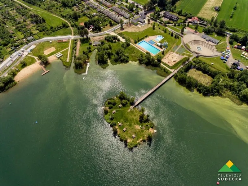 Widok z lotu ptaka na jezioro, otoczone zielenią, domkami, na środku jeziora wysepka.