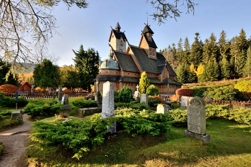 Zdjęcie małego, drewnianego kościółka  w otoczeniu zabytkowego cmentarza i przyrody