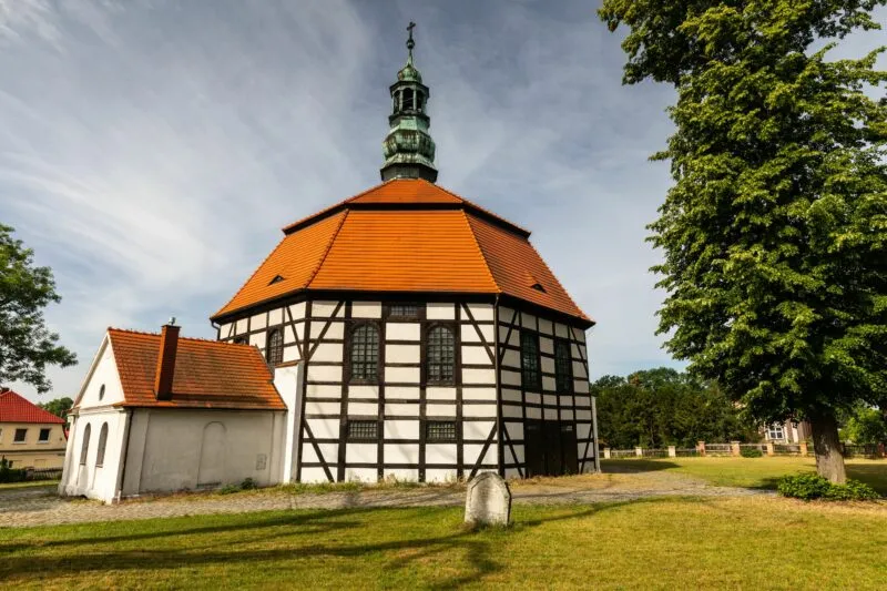 Zabytkowy kościół w Sułowie ma ryglową czarno-białą elewację, wysoki czerwony dach i małą wieżę, a wokół rozciąga się trawnik ze starymi drzewami.