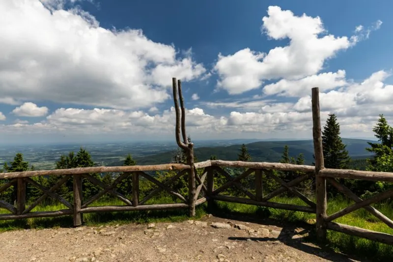 Drewniany taras widokowy na Stogu Izerskim otwiera szeroką panoramę doliny i odległych gór, widoczną między pniami, świerkami i białymi chmurami.