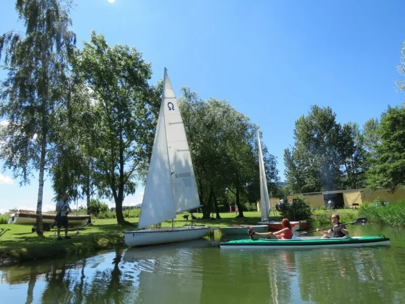 Widok na jezioro, na którym znajduje się kajak, łódki, otoczone drzewami.
