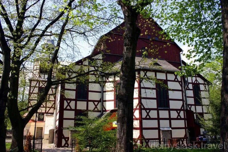 Hrazděné průčelí Kostela Míru v Jaworu s bílými poli a tmavými trámy se tyčí mezi zelenými stromy, částečně zakryté jarními větvemi.