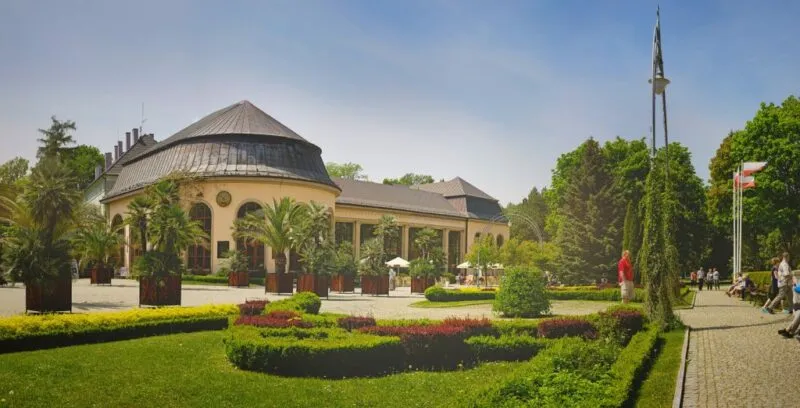 Budynek uzdrowiska z elegancką, historyczną fasadą, ozdobioną detalami w duchu architektury uzdrowiskowej, otoczony pięknym parkiem.