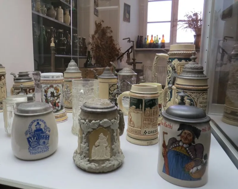 Stare zdobione malunkami kufle ustawione na białym stole. W oddali widać okno