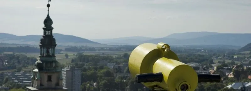 Z Twierdzy Kłodzko rozciąga się panorama miasta z kościelną wieżą, czerwonymi dachami i żółtą lunetą na pierwszym planie, na tle łagodnych gór.