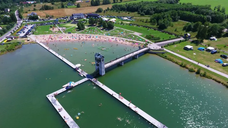Widok z lotu ptaka ukazuje Zalew w Starej Morawie z kąpieliskiem, pływającymi pomostami i wieżą, otoczony plażą, trawnikami, parkingami oraz polami.