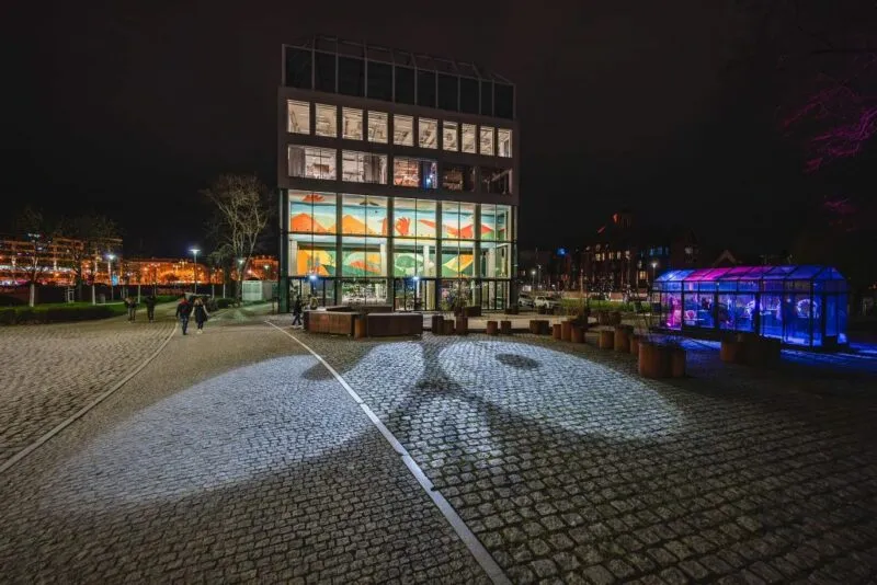 prostokątny budynek który jest restauracją. jest on cały przeszklony, zdjęcie zostało wykonane w nocy przez co widać wnętrze i w nim kolorowy mural znajdujące się w nim. oprócz budynku zdjęcie obejmuje rownież część wyspy słodowej we wrocławiu