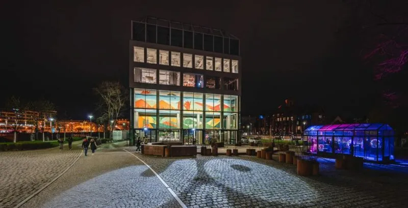 prostokątny budynek który jest restauracją. jest on cały przeszklony, zdjęcie zostało wykonane w nocy przez co widać wnętrze i w nim kolorowy mural znajdujące się w nim. oprócz budynku zdjęcie obejmuje rownież część wyspy słodowej we wrocławiu
