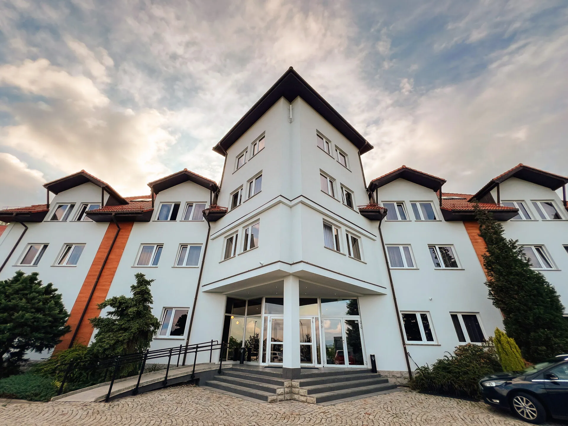 Widok na wejście główne do hotelu. Długi, trójkondygnacyjny budynek z wejściem wyszczególnionym symetrycznym, trójkątnym wykuszem.