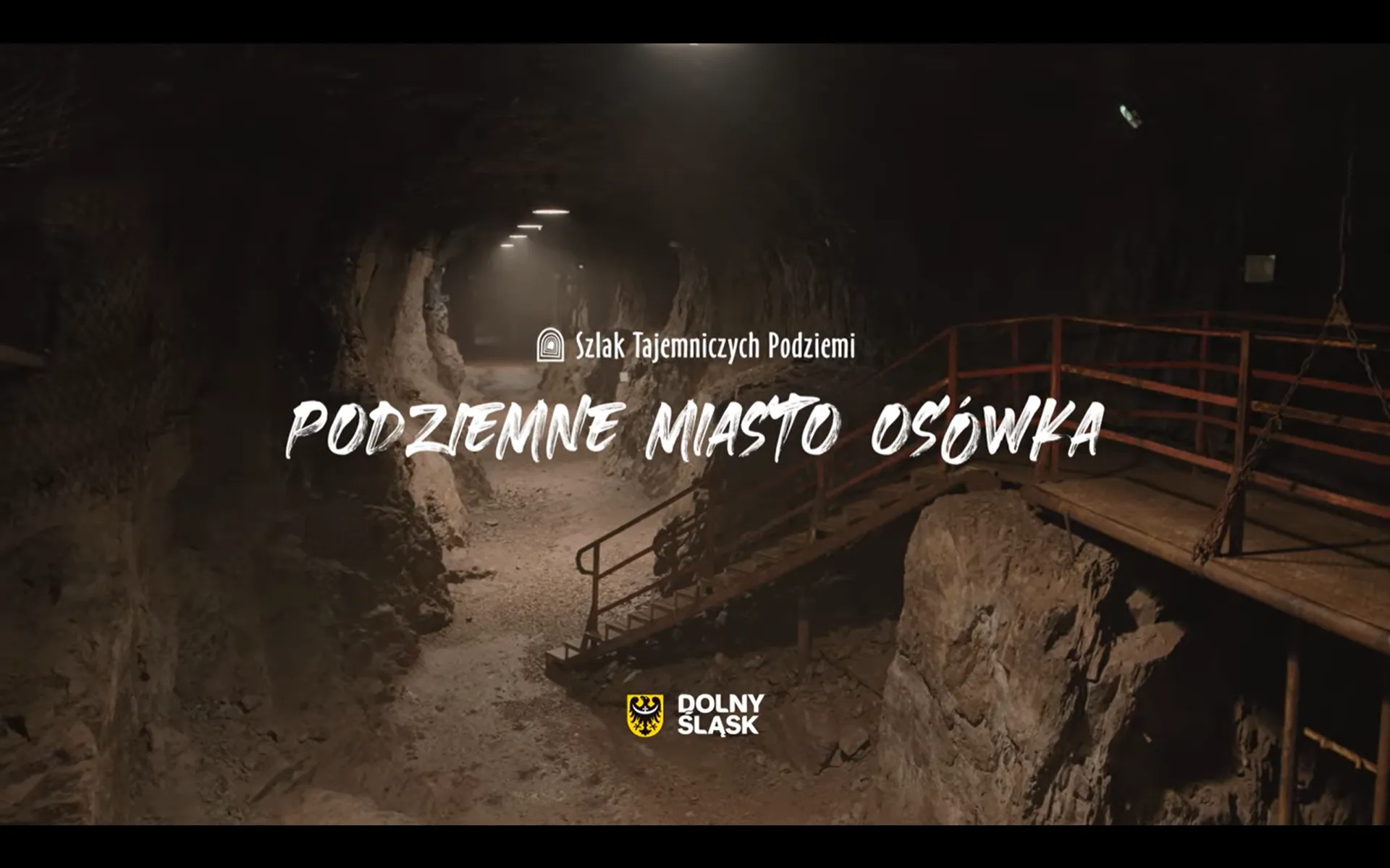 wysoki oraz długi wydrążony korytarz, lekko oświetlony. po prawej stronie znajdują się ciemno czerwone schody które prowadzą do wyżej położonego tunelu