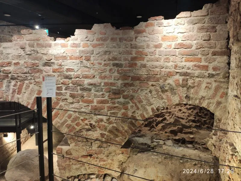 Drugie zdjęcie ukazuje fragment starego muru z cegły z łukiem, prawdopodobnie część fortyfikacji. Jest dobrze oświetlony i zabezpieczony barierkami.