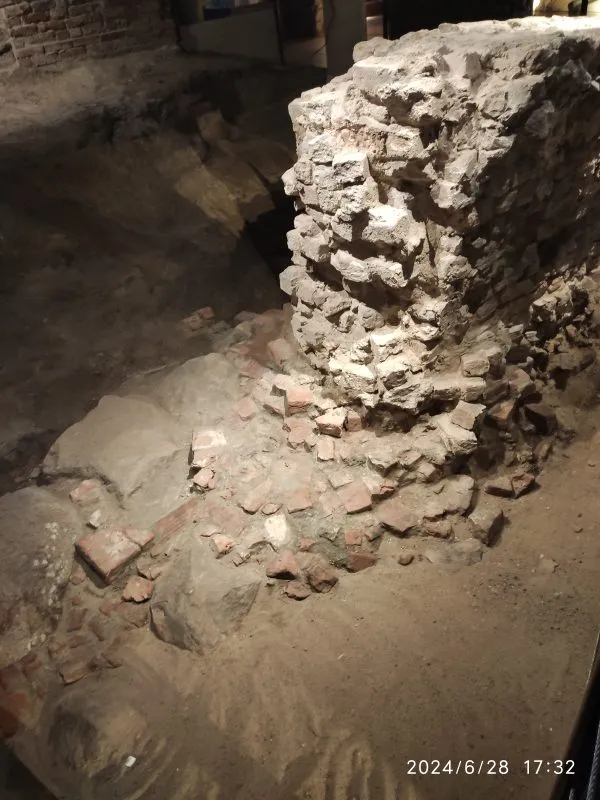 ruiny z cegieł i kamieni, częściowo zasypane piaskiem. To pozostałości dawnej konstrukcji odsłonięte podczas badań archeologicznych.
