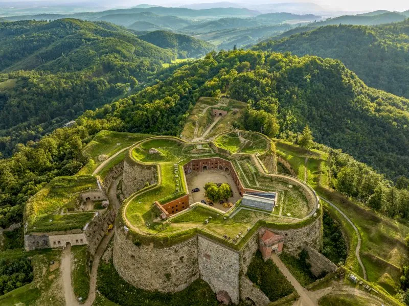 Majestatyczna twierdza górska na tle gór porośniętych gęstymi zielonymi lasami. 