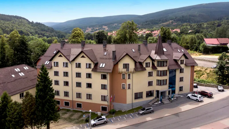 Zdjęcie z drona prezentujące duży, wielopiętrowy budynek hotelu. Przed obiektem znajduje się parking, wzdłuż ulicy, a na horyzoncie pasma górskie.
