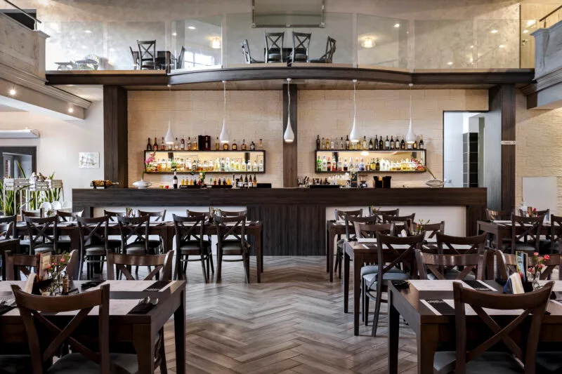 Sala restauracji z widocznym barem wykonanym w drewnie. Przed nim znajduje się kilkanaście drewnianych stolików z drewnianymi krzesłami.
