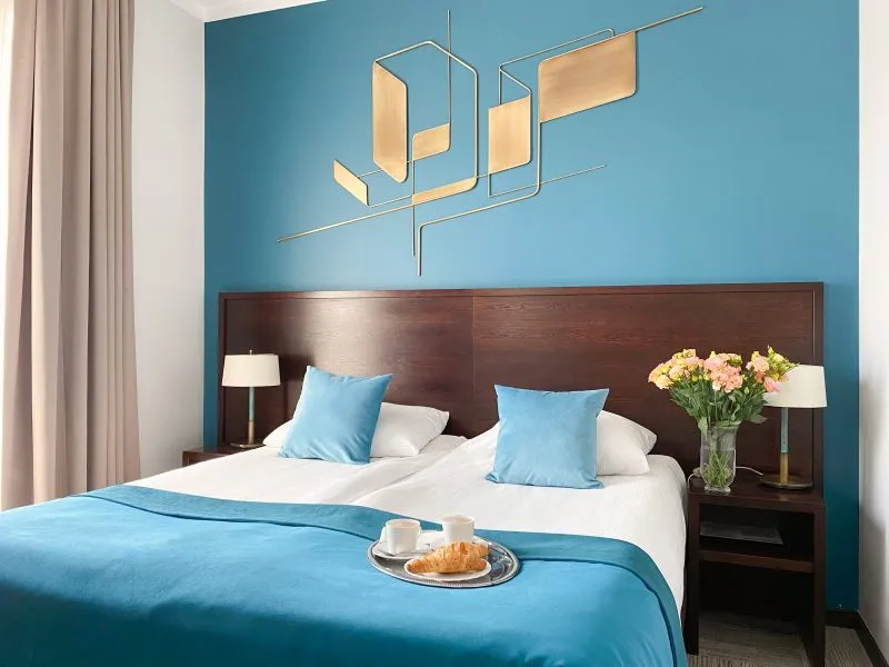 Zdjęcie przedstawia nowoczesny, elegancki pokój hotelowy urządzony w minimalistycznej stylistyce. Dominują w nim neutralne kolory – biel, turkus i drewno. W pokoju znajdują się dwa pojedyncze łóżka ustawione obok siebie, z drewnianym zagłówkiem. Na łóżkach leżą małe turkusowe poduszki i narzuta w tym kolorze oraz poczęstunek powitalny.