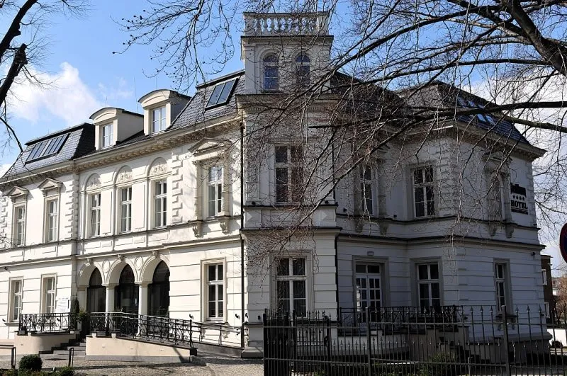 Elegancki, zabytkowy budynek w klasycystycznym stylu, w którym mieści się hotel Red Baron w Świdnicy. Jasna elewacja ozdobiona jest licznymi zdobieniami, a duże okna i mansardowy dach z nowoczesnymi świetlikami nadają mu wyjątkowy charakter. Przed wejściem znajduje się stylowe, czarne ogrodzenie oraz schody prowadzące do głównych drzwi hotelu.