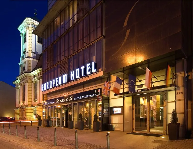 Zdjęcie nocne. Dobrze oświetlona, nowoczesna szklana fasada hotelu Europeum kontrastuje z zabytkowym sąsiedztwem, w tym z barokowym kościołem po lewej stronie. Budynek wyróżnia się minimalistycznym designem, dużymi przeszkleniami i eleganckim, geometrycznym wykończeniem.