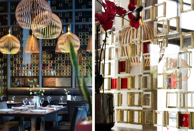 Wnętrze restauracji hotelowej charakteryzuje się eleganckim i nowoczesnym wystrojem z designerskimi lampami i geometrycznymi dekoracjami na ścianach. Ciepłe oświetlenie i starannie ułożone nakrycia na stołach tworzą przytulną atmosferę. Szklana przegroda z elementami w odcieniach złota i czerwieni dodaje wnętrzu luksusowego charakteru.
