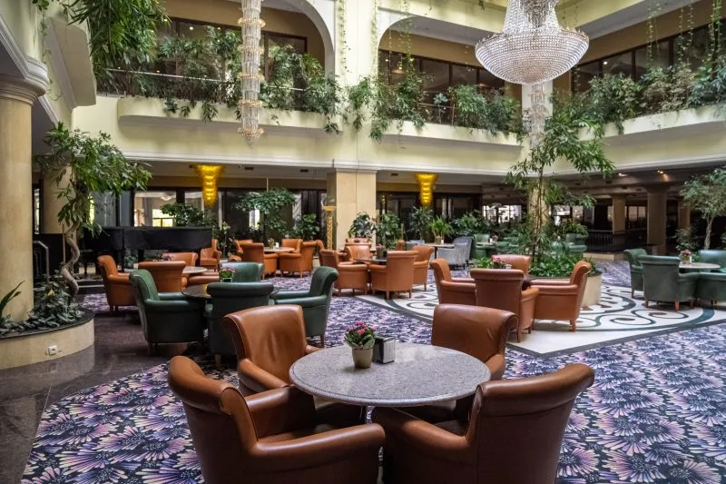 Patio hotelowe. Na podłodze ozdobne płytki. Kilkanaście okrągłych stolików z masywnymi skórzanymi fotelami. Z balkonów wewnętrznych zwisa duża ilość roślin zielonych. 
